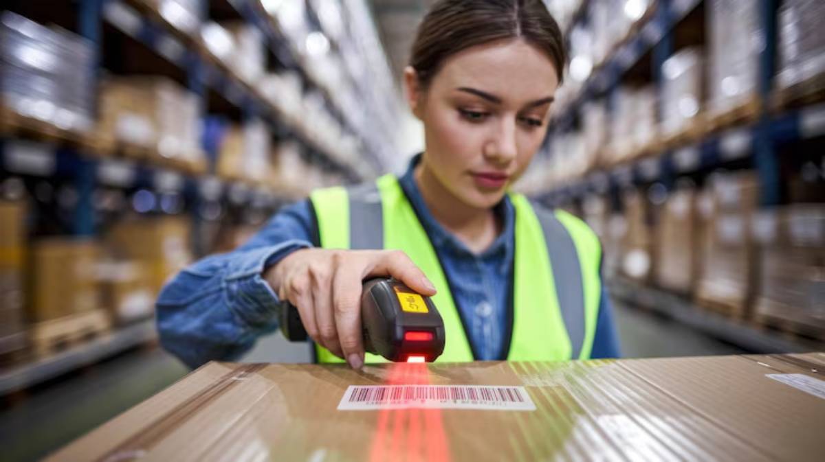 A warehouse worker scans a barcode on a cardboard box using a handheld scanner, surrounded by shelves filled with packages.