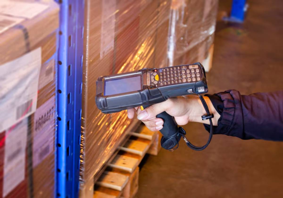 A worker holds a handheld barcode scanner, poised to scan packages stacked on a pallet in a warehouse setting.