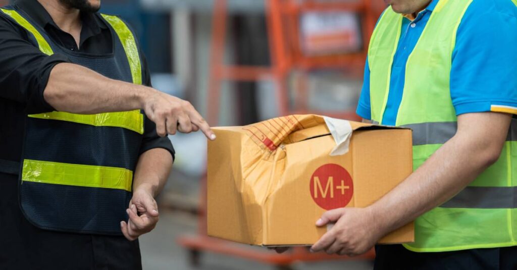 Two workers in safety vests discuss a damaged package labeled M while handling it in a warehouse environment.