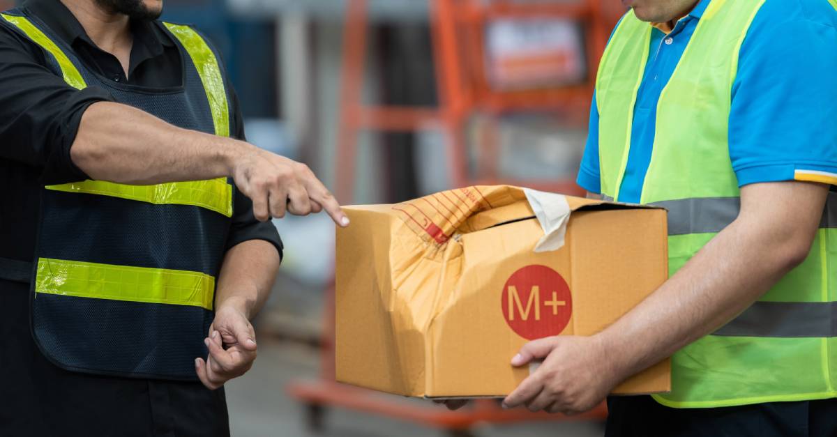 Two workers in safety vests discuss a damaged package labeled M while handling it in a warehouse environment.