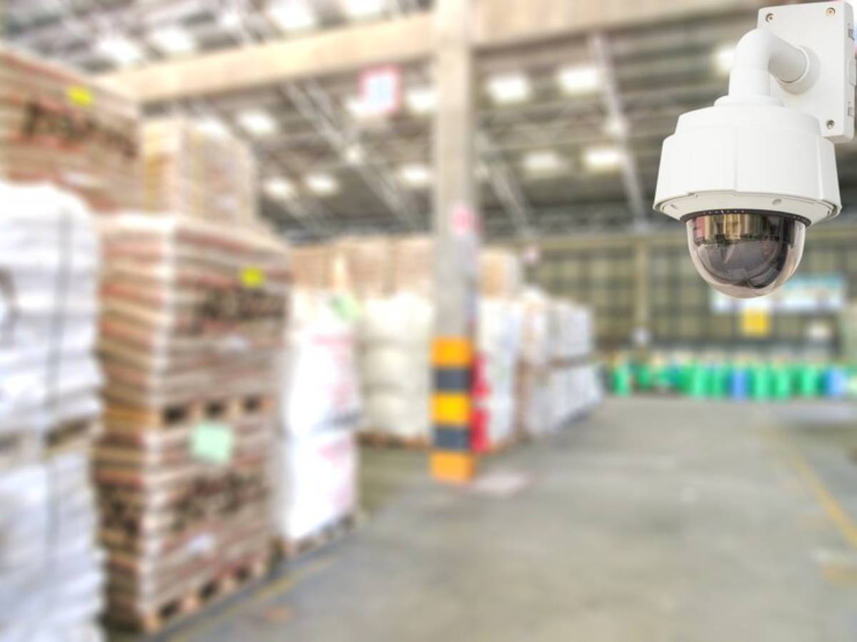 A surveillance security camera mounted on a wall, overlooking pallets of goods in a large warehouse space.
