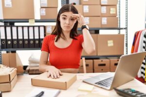 A woman in a red shirt sits at a desk with a laptop, surrounded by cardboard boxes and storage files, appearing thoughtful.