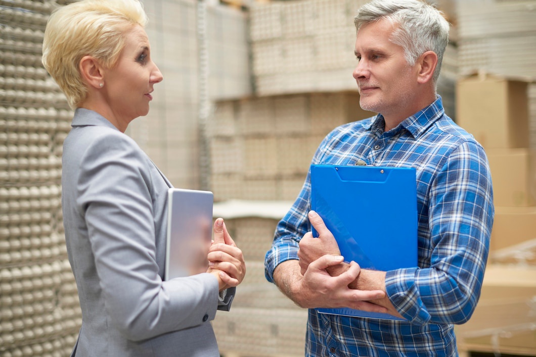 A professional conversation between two people in a warehouse, one holding a tablet and the other a blue clipboard.