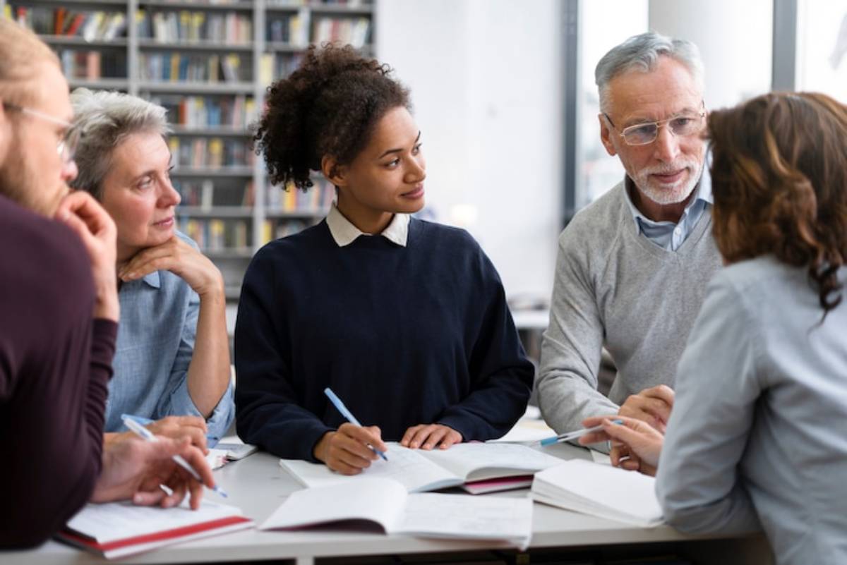 A diverse group of individuals engaged in a collaborative discussion at a table filled with books and notebooks, in a bright library setting.