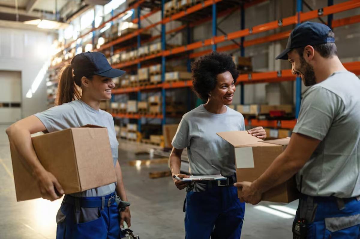 Three warehouse workers in gray shirts and blue pants are collaborating, handling boxes amid shelves stocked with goods.