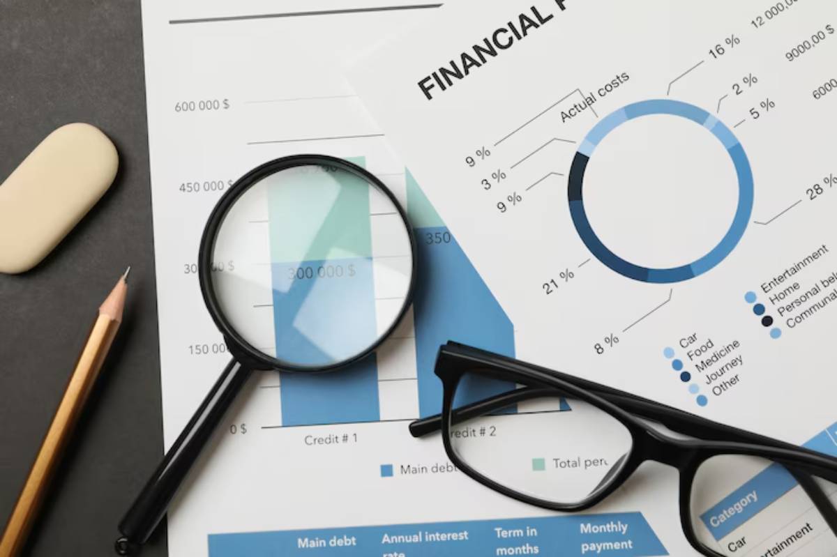 A magnifying glass and glasses rest atop financial reports displaying charts, graphs, and data about costs and debt.