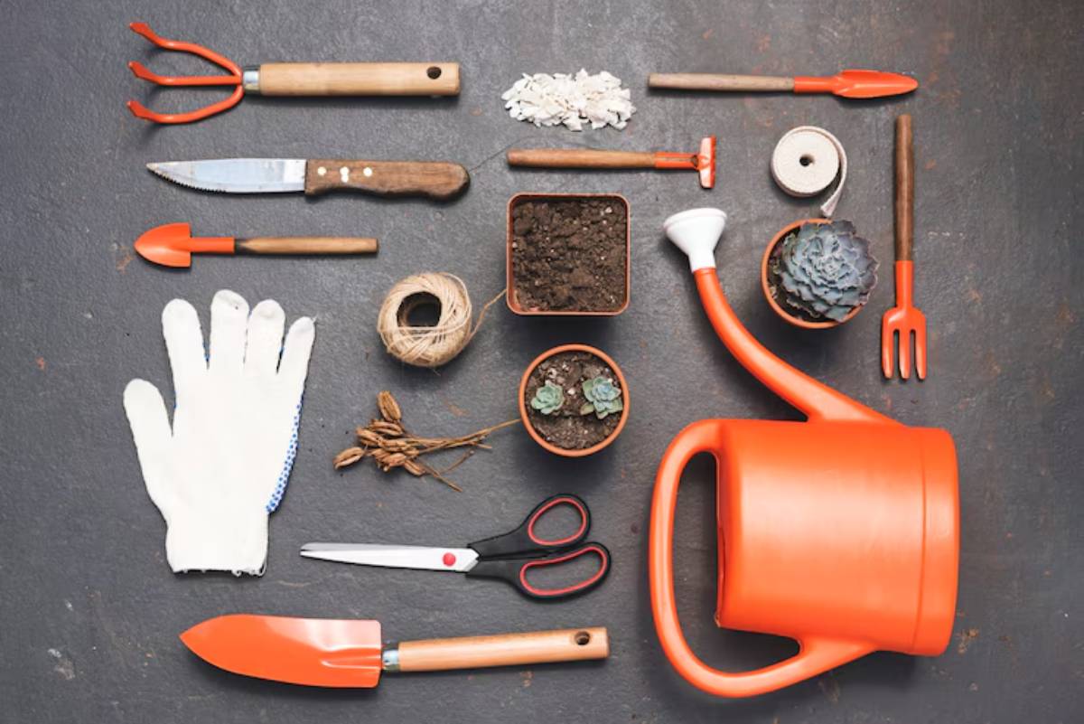 A flat lay of gardening tools including a watering can, pots, gloves, a knife, and various hand tools on a dark surface.