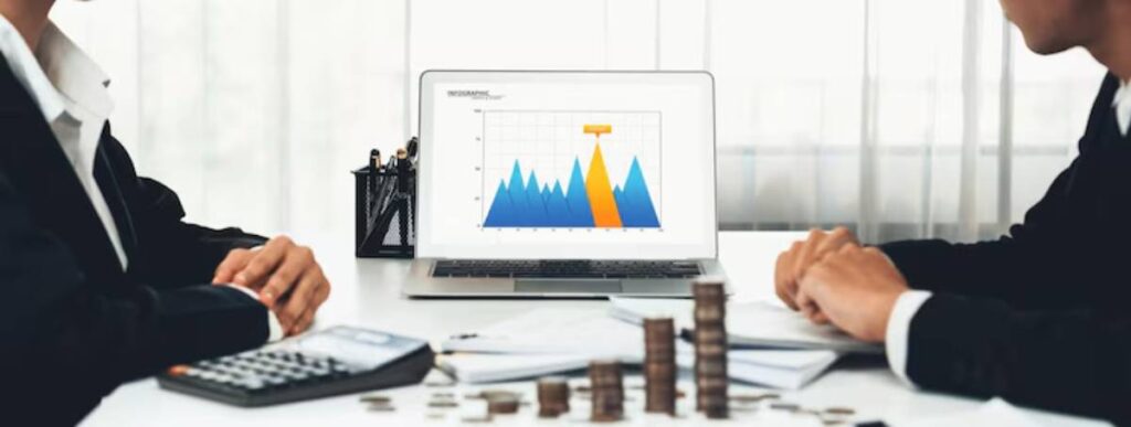 Two professionals discuss financial data, with a laptop displaying a colorful graph and stacks of coins on the table.