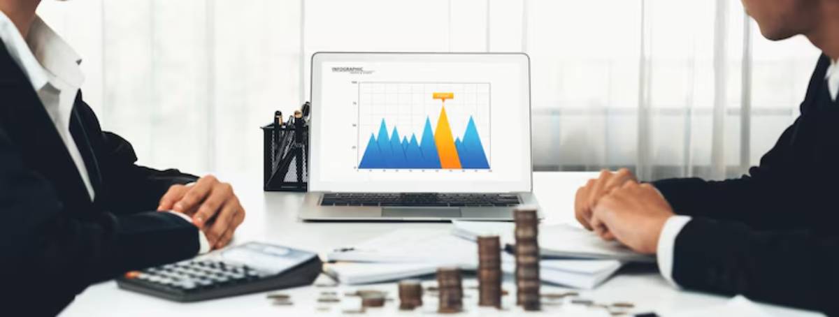 Two professionals discuss financial data, with a laptop displaying a colorful graph and stacks of coins on the table.