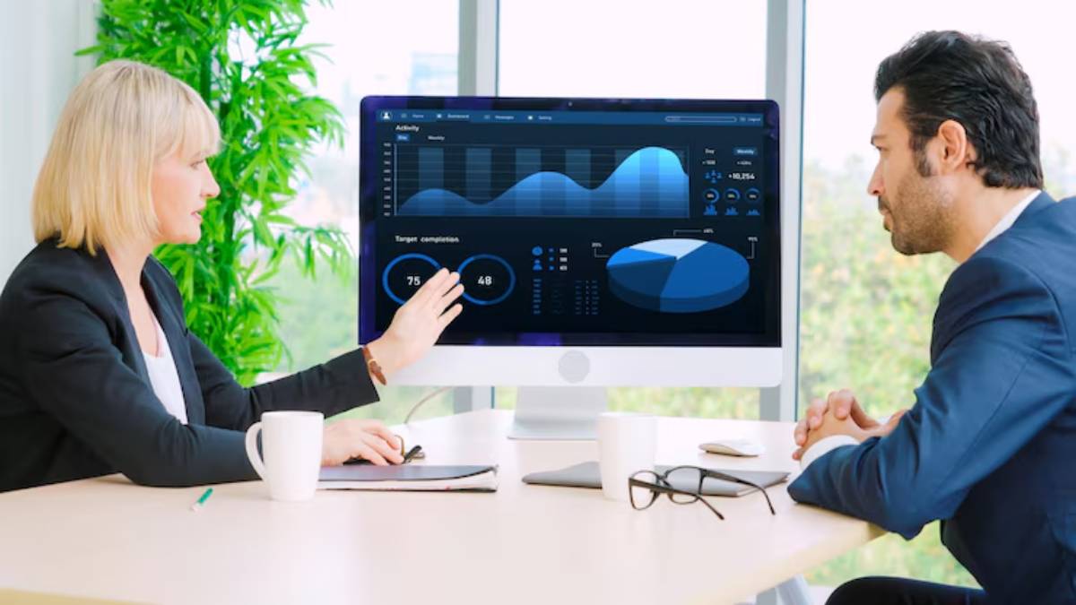 A business meeting with a woman pointing at data visualizations on a desktop monitor while a man observes attentively.