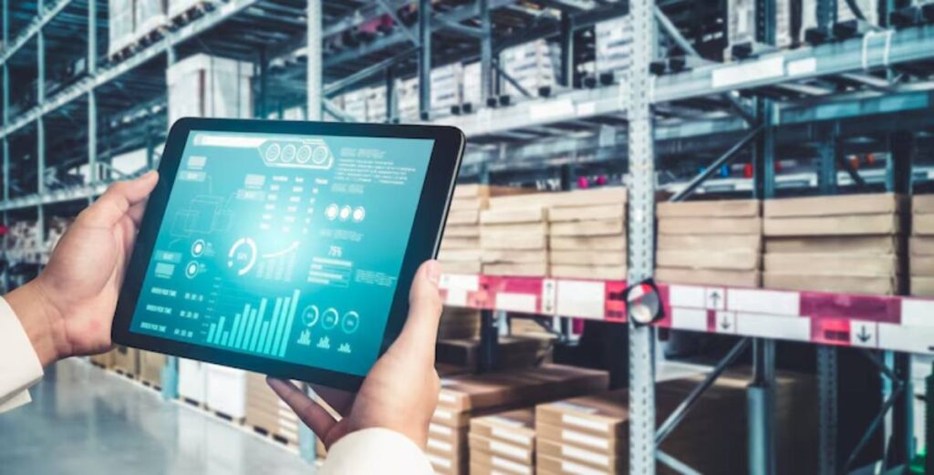 A person holds a tablet displaying inventory data in a warehouse filled with stacked boxes on shelves.