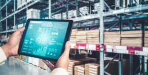 A person holds a tablet displaying inventory data in a warehouse filled with stacked boxes on shelves.