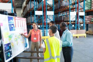 A group of four professionals discusses a world map and data charts in a warehouse setting, focusing on logistics and analysis.