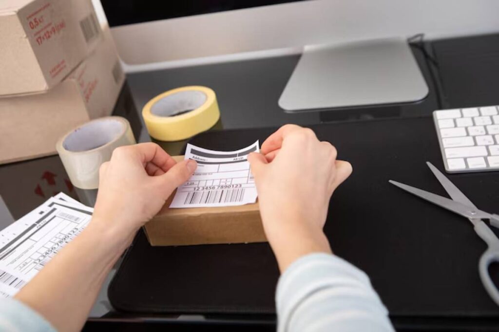 A person attaches a shipping label on a cardboard box at a desk, surrounded by tape, scissors, and a computer monitor.