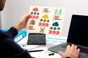 A person holding a colorful infographic while using a laptop and calculator at a desk with various printed materials.