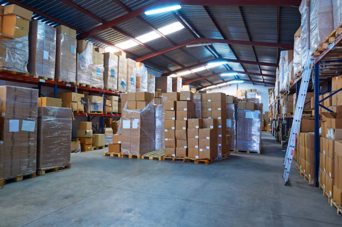 A large warehouse with tall shelves filled with stacked cardboard boxes, some wrapped in plastic, and a metal ladder against the wall.