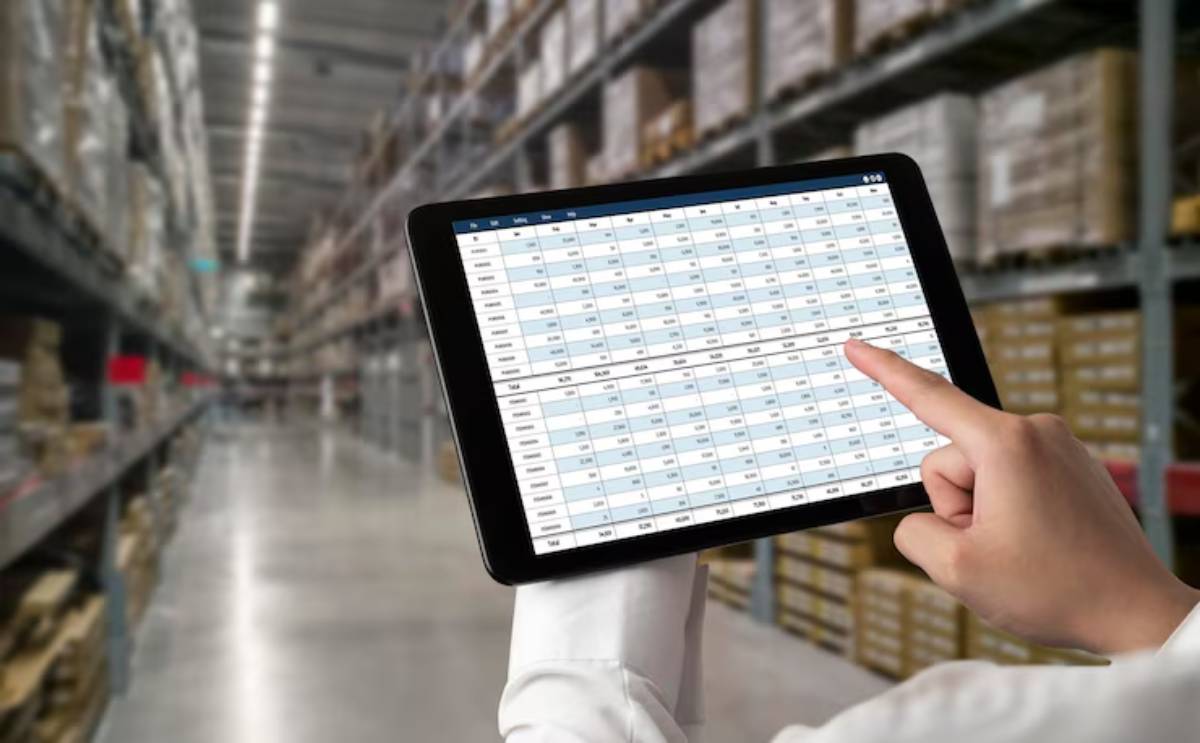 A person in a white shirt points at a digital tablet displaying a data spreadsheet in a spacious warehouse filled with boxes and shelves.