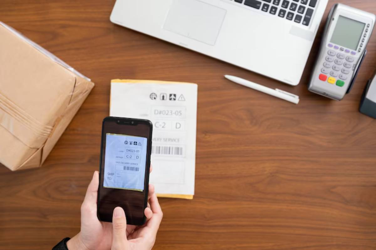 A person scans a shipping label on their phone, with a package, laptop, pen, and card reader on a wooden desk.