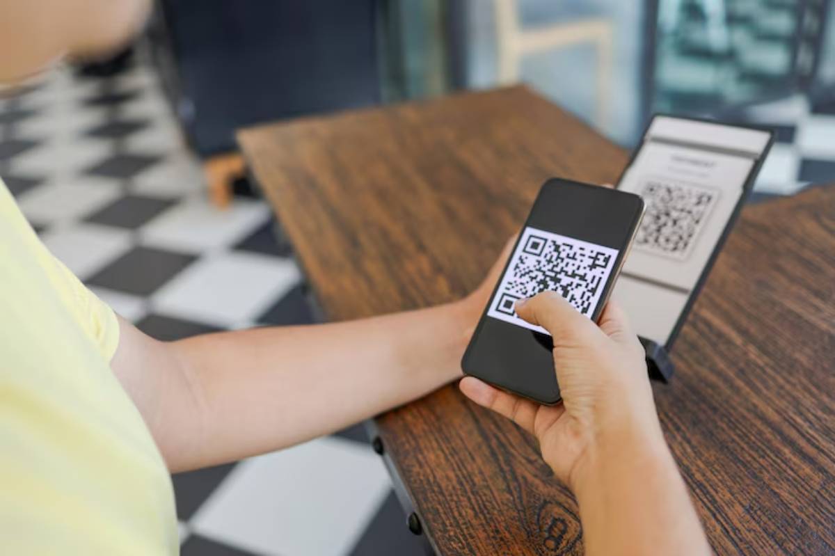 A person scans a QR code with a smartphone at a table, while a menu with another QR code is displayed beside them.