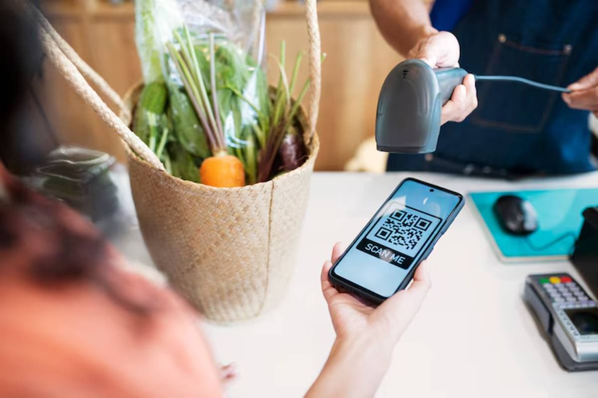 A shopper holds a smartphone displaying a QR code while a cashier scans items from a basket filled with fresh vegetables.