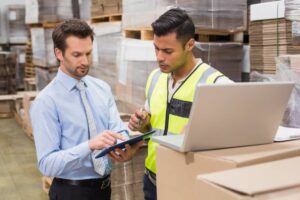 Two men discuss logistics in a warehouse, one holding a tablet and the other holding a pen, with boxes and stacks of material around them.