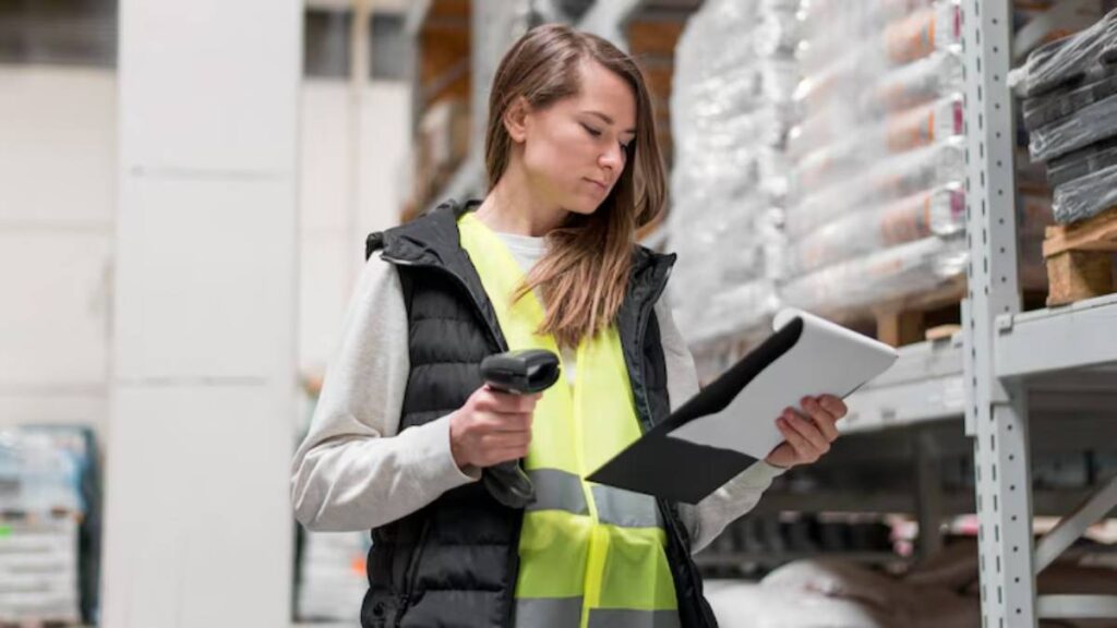 A woman in a safety vest scans items with a barcode scanner while reviewing a clipboard in a warehouse filled with stacked goods.