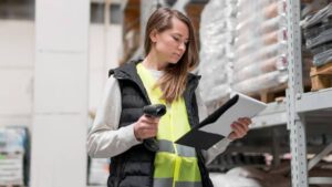 A woman in a safety vest scans items with a barcode scanner while reviewing a clipboard in a warehouse filled with stacked goods.