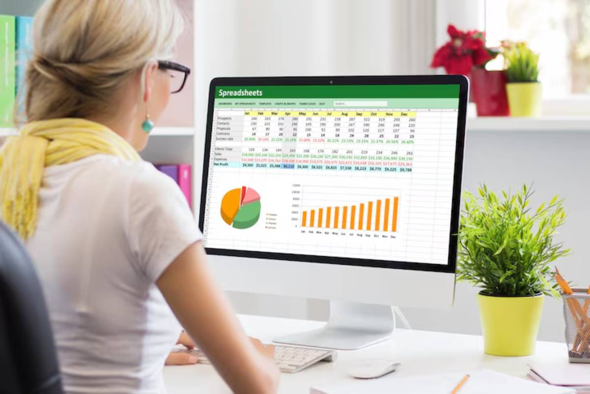 A woman with a yellow scarf analyzes financial data on a computer screen displaying spreadsheets and charts, surrounded by plants and books.