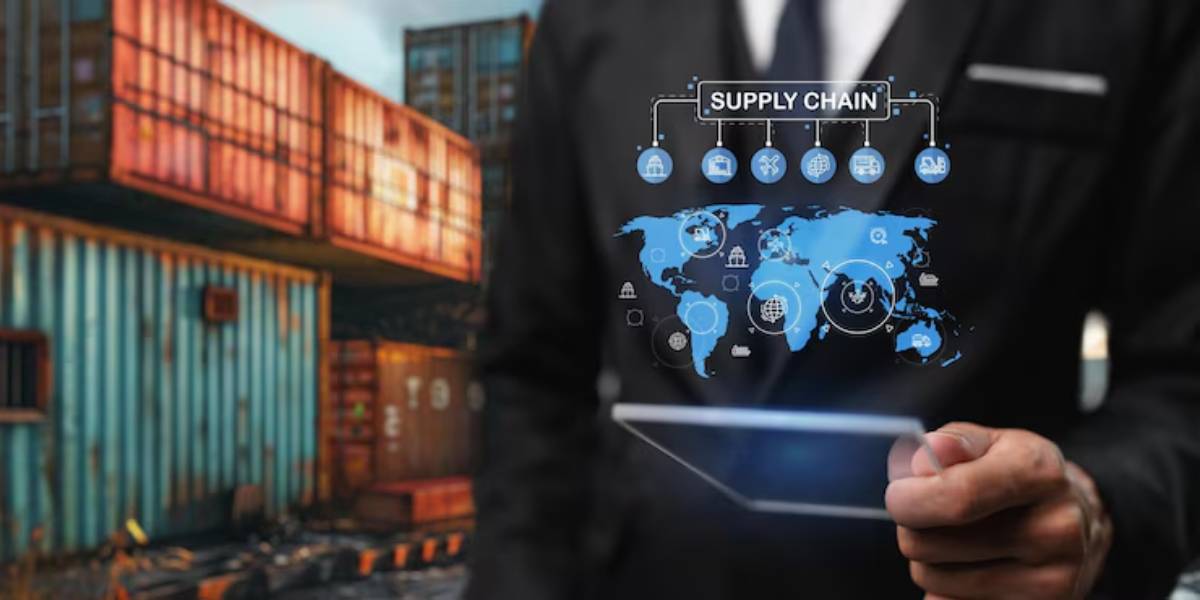 A businessman in a suit holds a tablet, with a digital supply chain graphic overlay and shipping containers in the background.