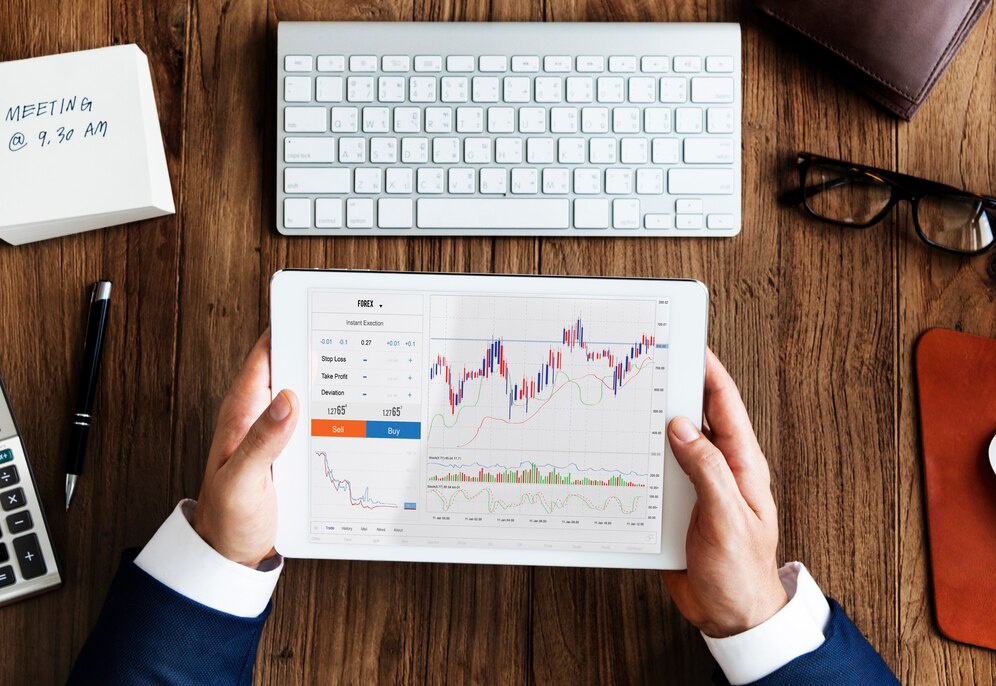 A person in a suit holds a tablet displaying a forex trading interface, surrounded by office supplies and a keyboard on a wooden desk.