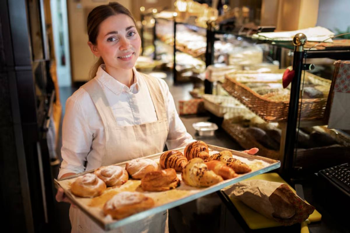 A bakery employee in a white apron holds a tray of freshly baked pastries, including cinnamon rolls and croissants, in a warm setting.