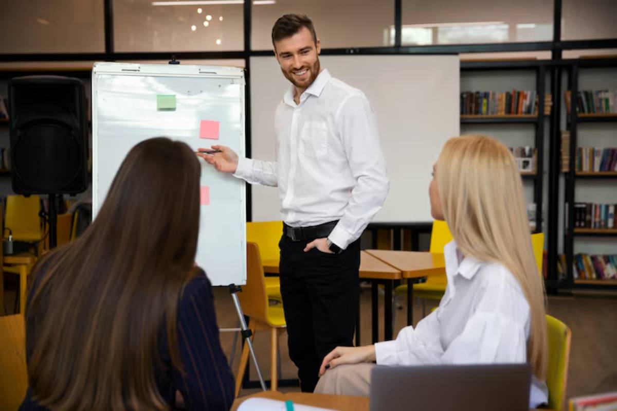 A presentation in a modern office setting, featuring a presenter by a flip chart and attentive audience members seated at tables.