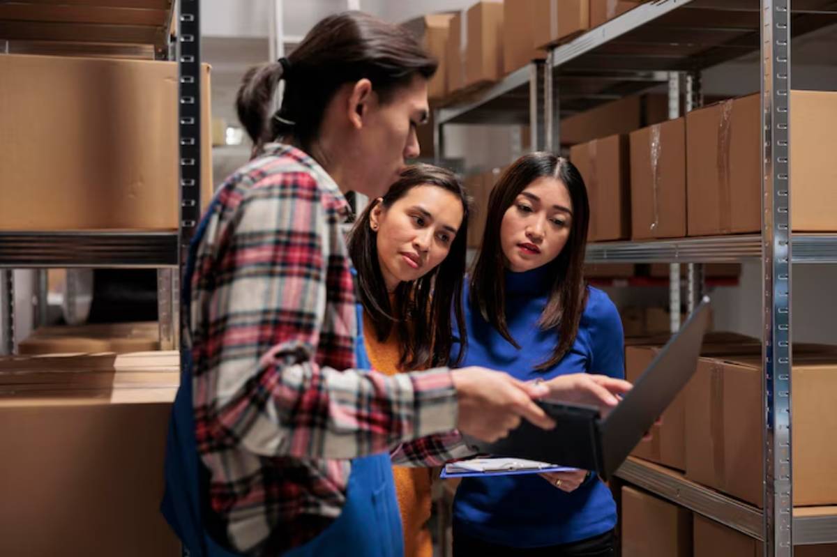 A person in a flannel shirt discusses details with two women in a warehouse filled with stacked cardboard boxes, holding a laptop and clipboard.