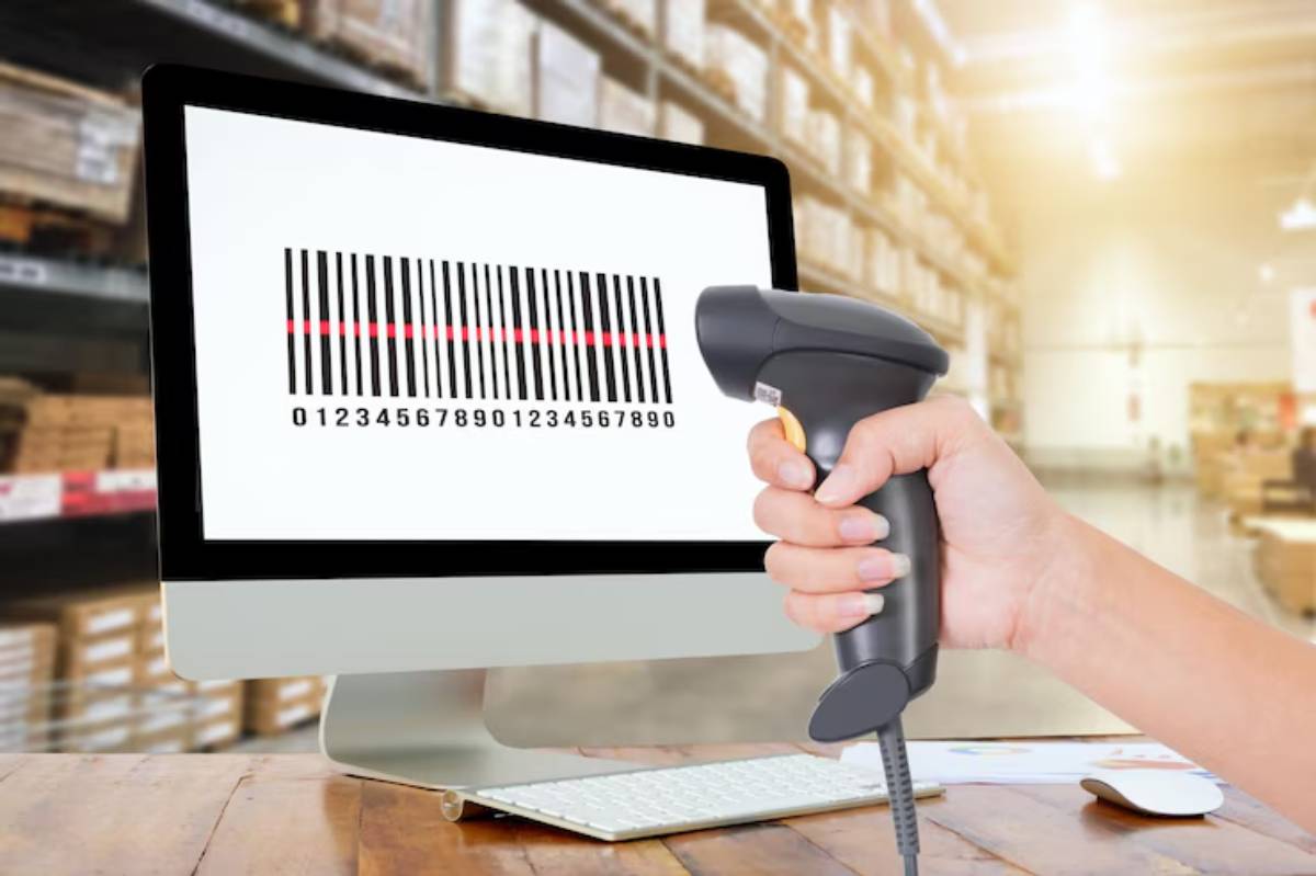 A hand holds a barcode scanner aimed at a computer screen displaying a barcode, set in a warehouse environment.