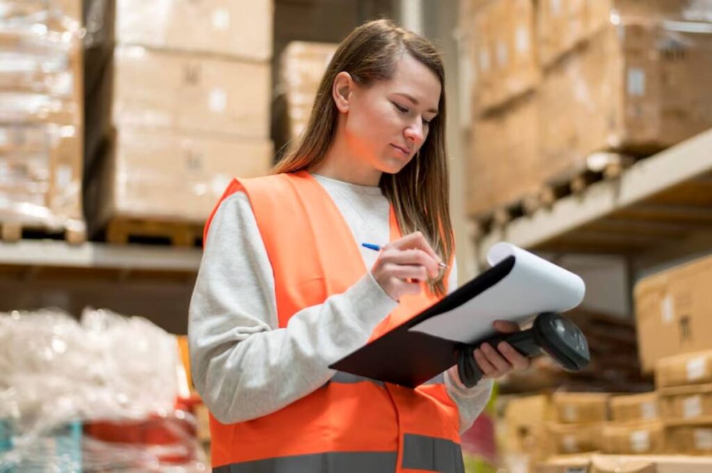 A woman in an orange safety vest stands in a warehouse, holding a clipboard and making notes amidst stacks of boxes.