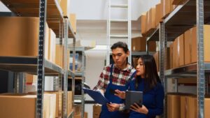 Two workers in a warehouse discuss inventory while holding a clipboard and a tablet, surrounded by shelves of cardboard boxes.