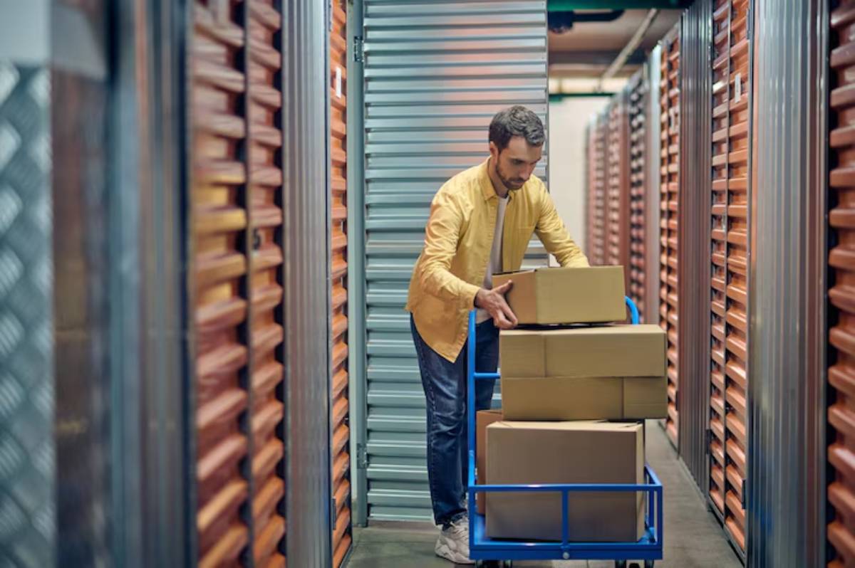 A person in a yellow shirt pushes a cart loaded with boxes down a storage facility aisle, flanked by metal storage doors.