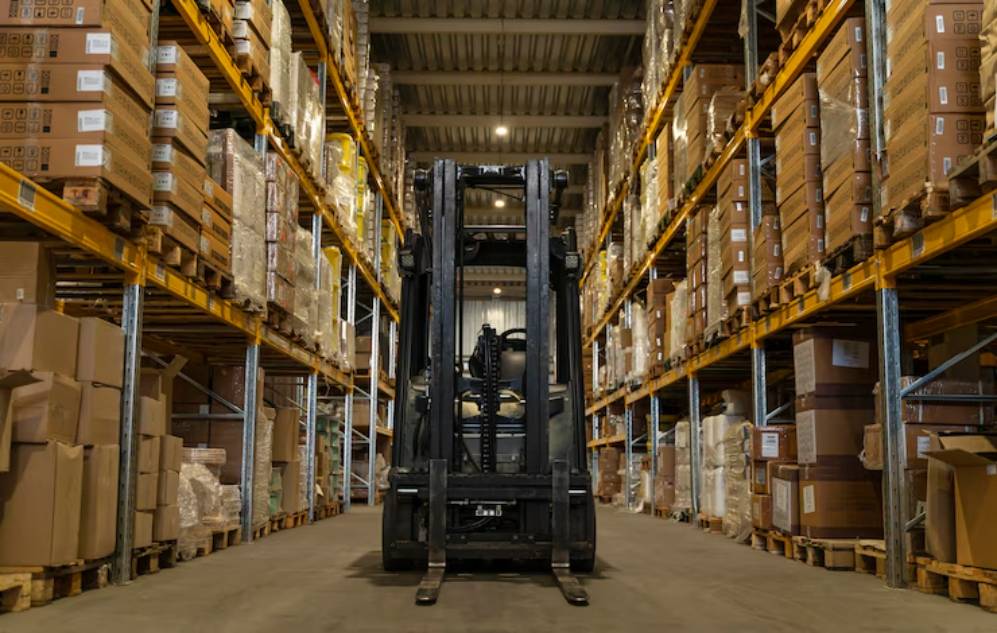 A view of a warehouse with a black forklift centered on the aisle, surrounded by tall shelves stacked with cardboard boxes.