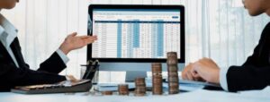 A person points at a spreadsheet on a computer screen, while another examines financial data and stacks of coins on the table.