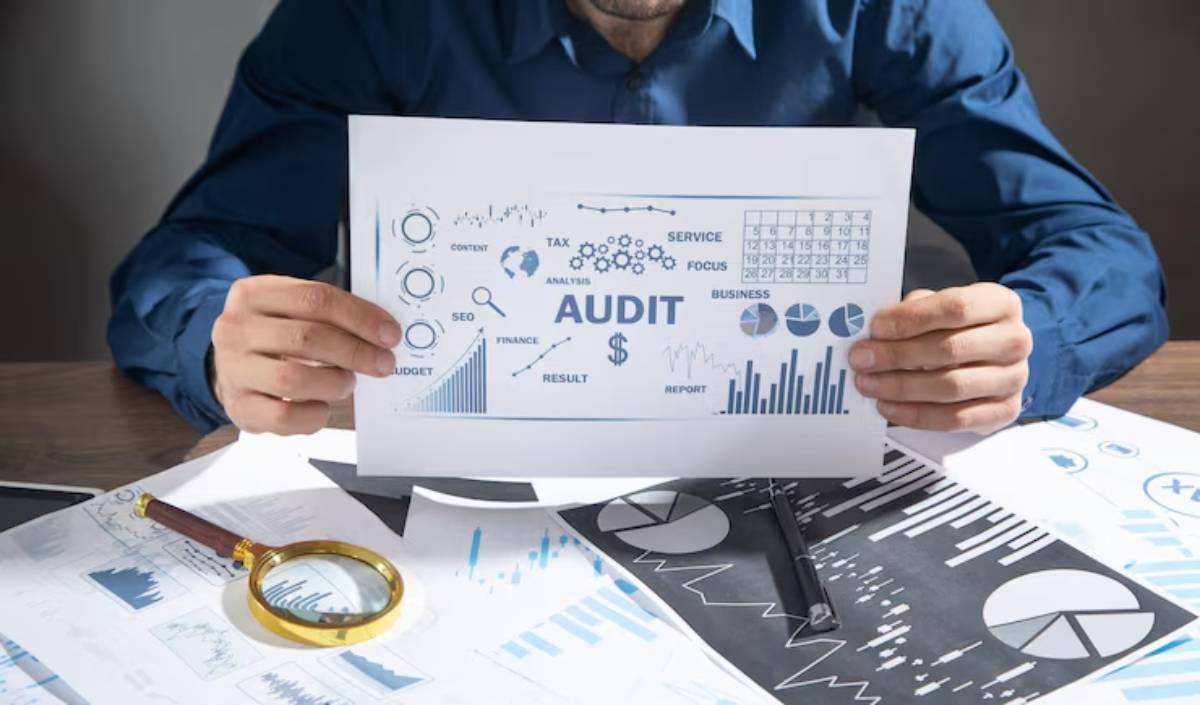 A person in a blue shirt holds an AUDIT report, surrounded by papers featuring graphs, charts, and analysis visuals.