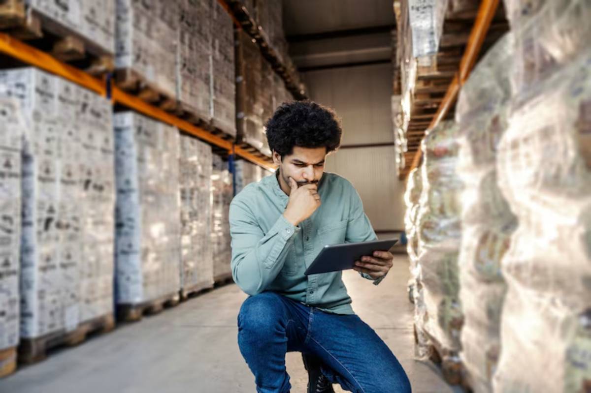 A warehouse worker in a blue uniform holds a clipboard, surrounded by shelves stocked with various products.
