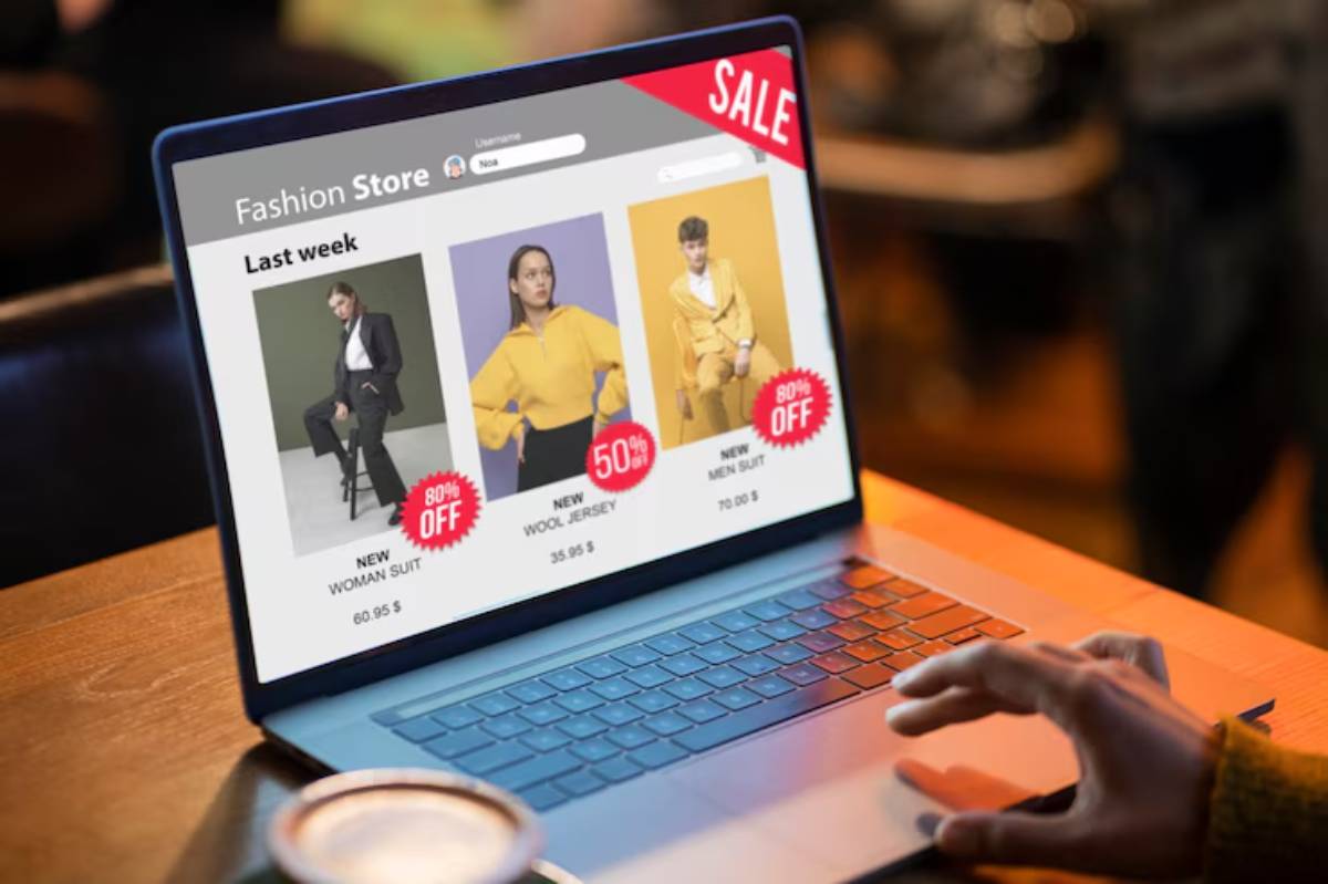 A hand interacts with a laptop displaying a fashion store website featuring discounted women’s and men’s suits.