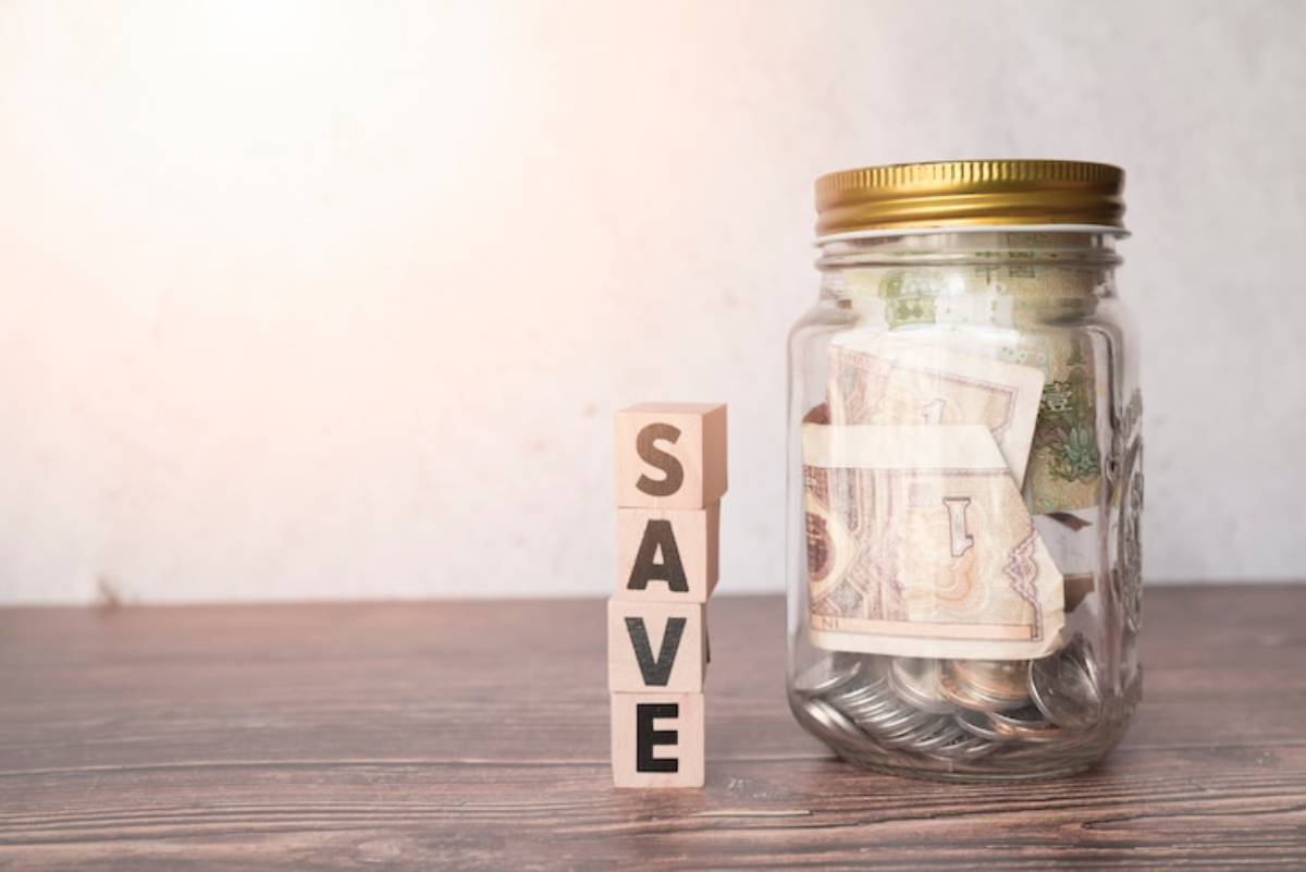 A glass jar filled with currency notes and coins beside wooden blocks spelling SAVE on a wooden surface.