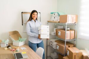 A person stands in an organized workspace holding several stacked boxes, with shelves of packed items and office supplies in the background.