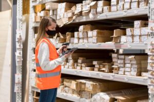 A warehouse worker in an orange vest scans inventory on a handheld device, surrounded by neatly stacked packages on shelves.