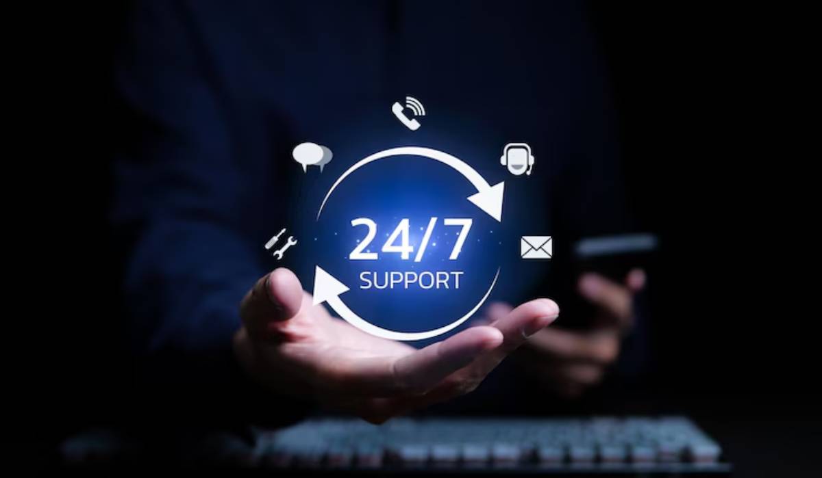 A hand holding a glowing icon displaying 24/7 Support, surrounded by communication symbols like phone, chat, email, and headset.