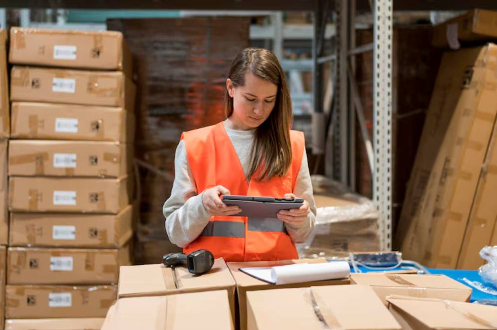 A warehouse worker in an orange safety vest uses a tablet while surrounded by cardboard boxes and packing supplies.