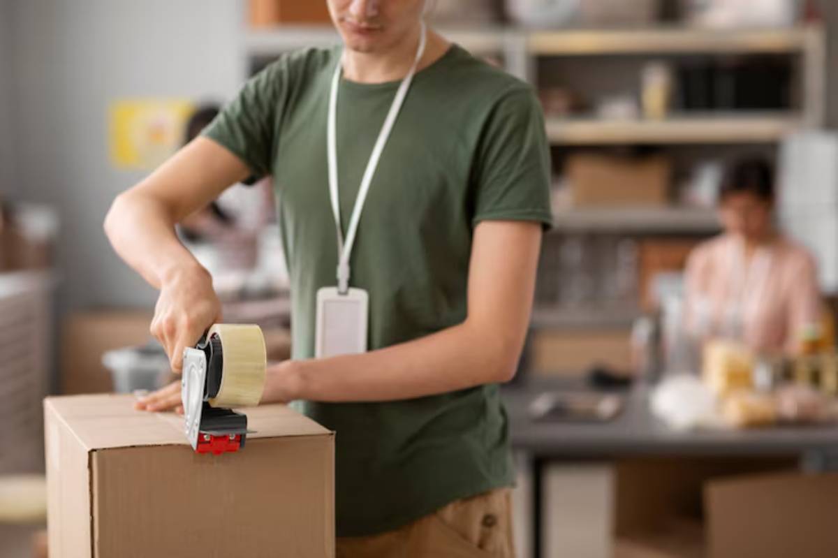 A person in a green shirt uses tape to seal a cardboard box in a busy workspace filled with shelves and supplies.