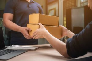 A delivery person hands over two stacked cardboard boxes to a person sitting at a desk with a laptop and paperwork.