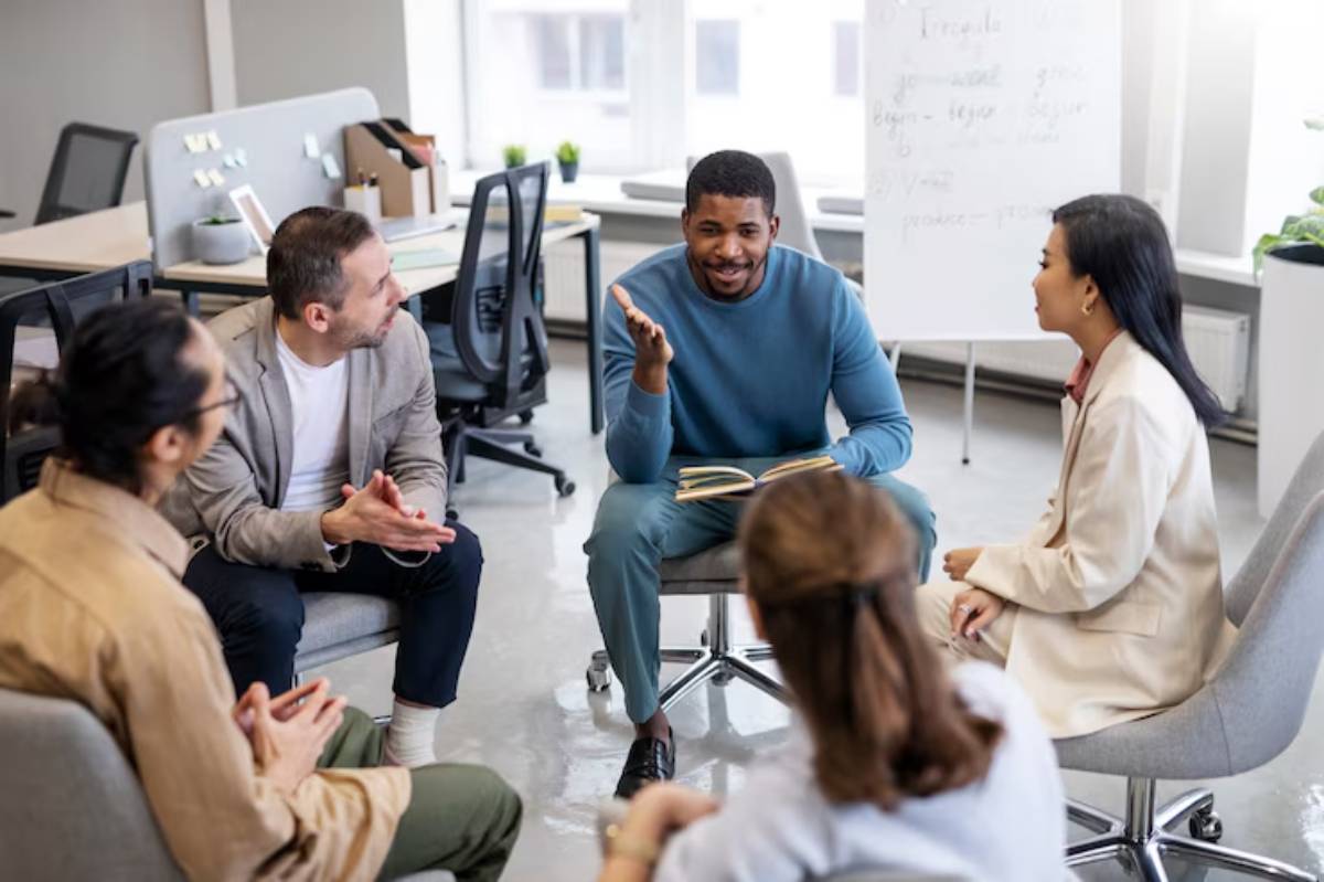 A group of five professionals sit in a modern office, engaged in a discussion, with one person holding a book and gesturing while speaking.
